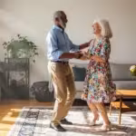 A happy senior couple dances energetically together in their sunlit living room, smiling and full of life.